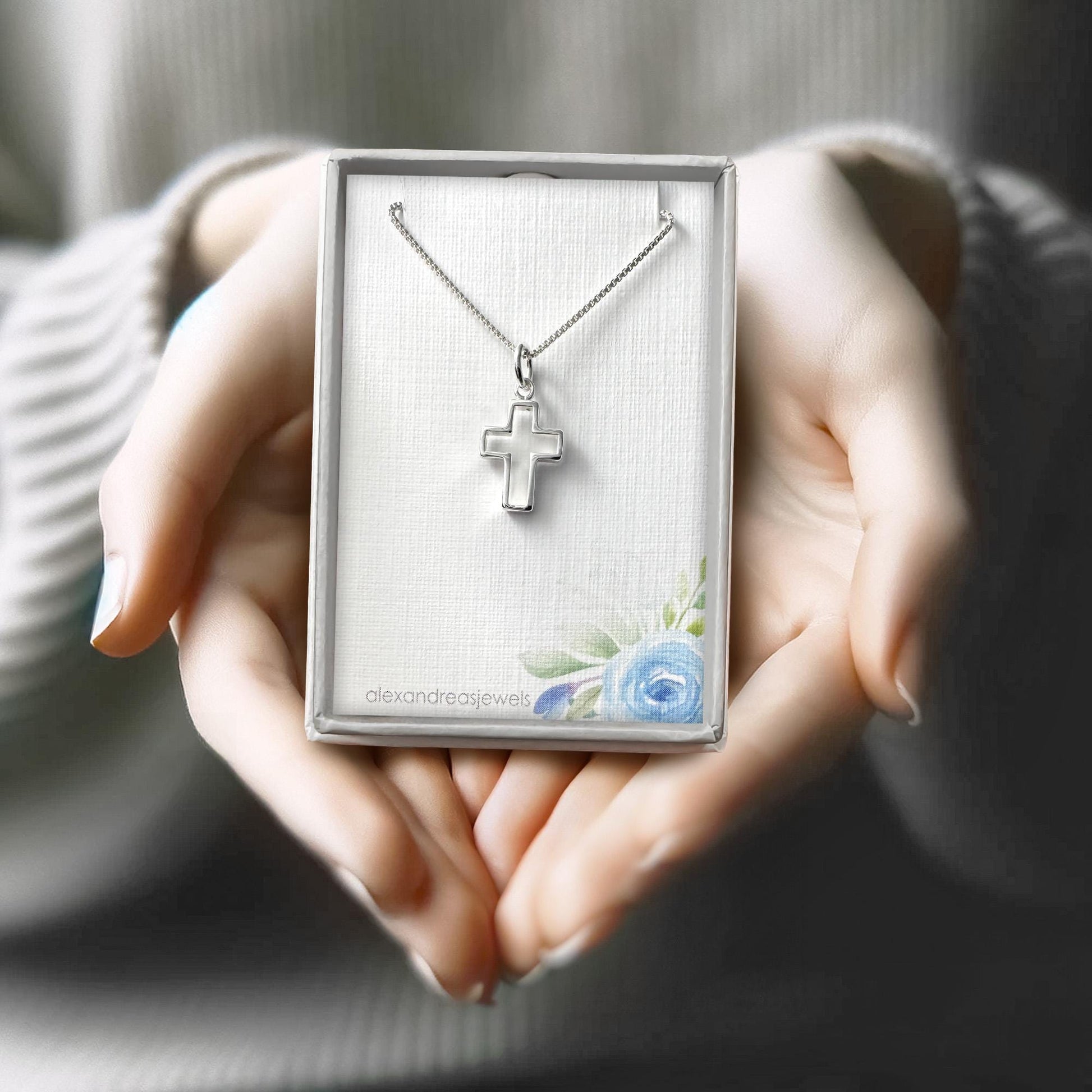 A person's hands holding a silver cross necklace in a box with a floral design.