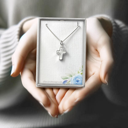 A person's hands holding a silver cross necklace in a box with a floral design.