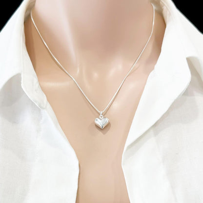 A silver heart-shaped pendant necklace is displayed on a mannequin's neck, wearing a white collared shirt.