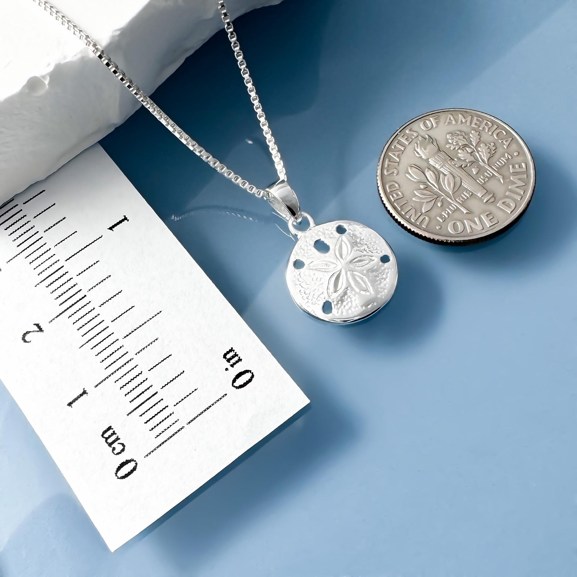 A silver necklace with a pendant is placed next to a U.S. one dollar coin on a blue surface.