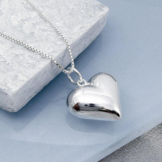 A silver heart-shaped pendant necklace is displayed on a gray surface, with a gray stone or block in the background.