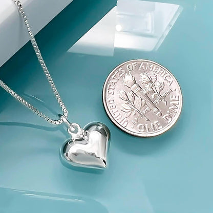 A silver heart-shaped pendant necklace and a United States one dollar coin are placed on a blue surface.