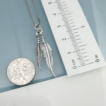a silver feather pendant necklace and a United States quarter coin, both resting on a white surface.