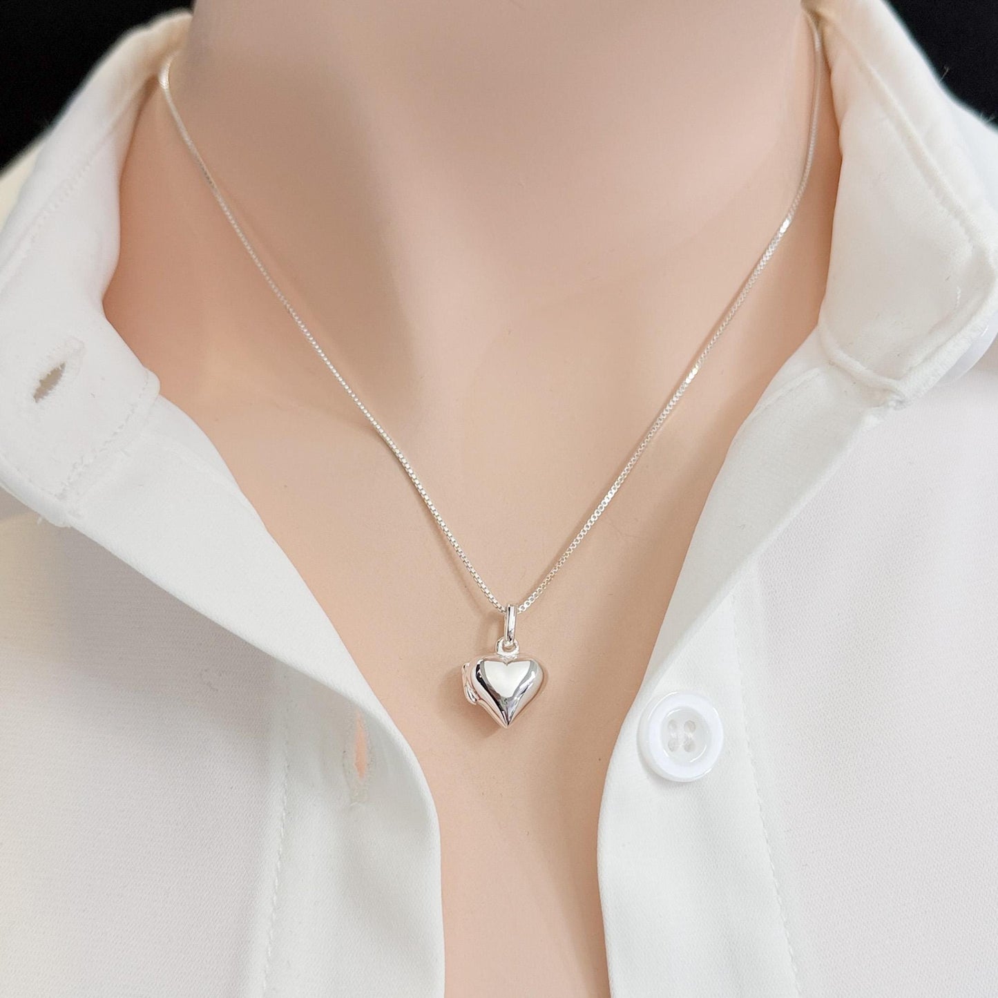 A silver heart-shaped pendant necklace is worn around a person's neck, with a white collared shirt visible underneath.