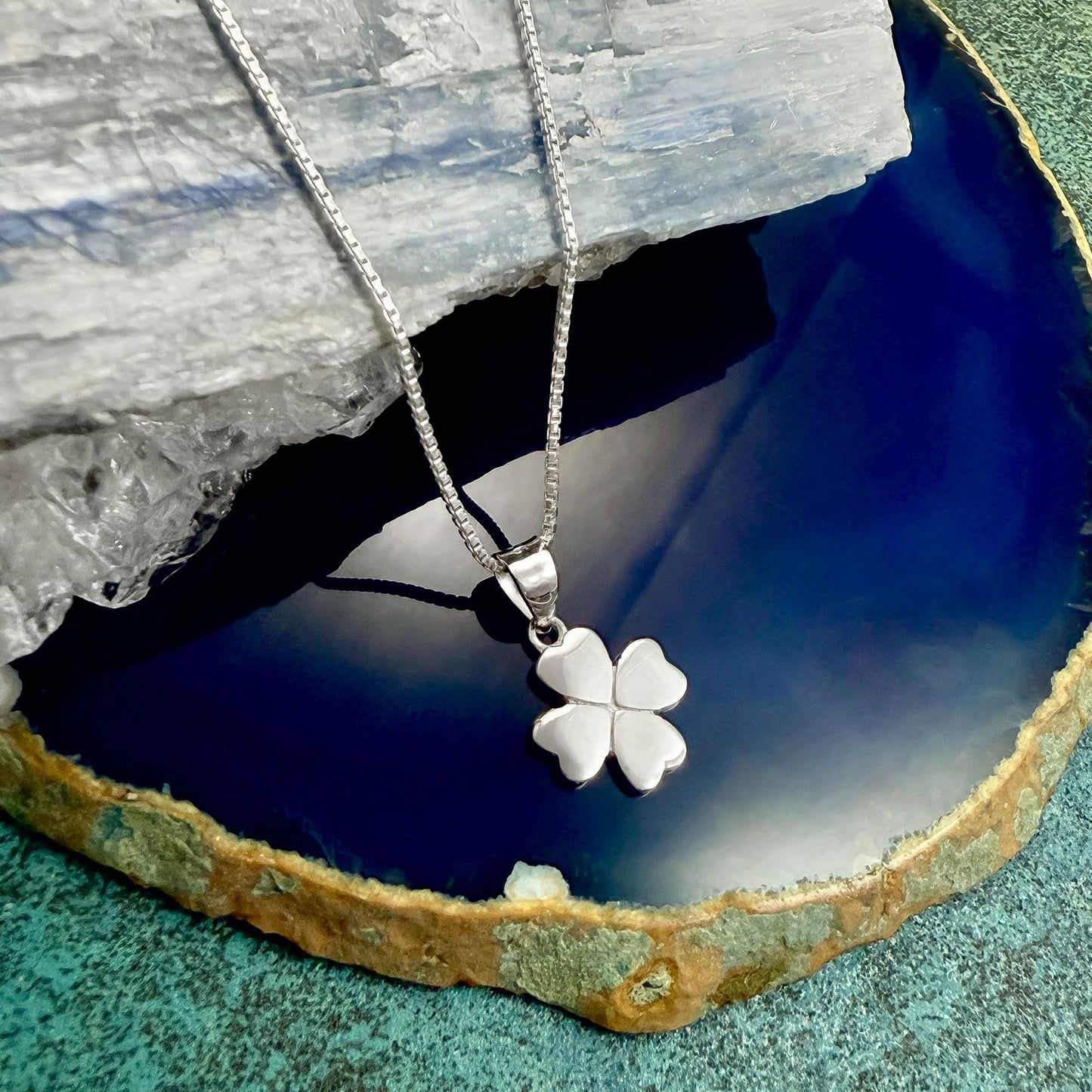A silver-colored pendant with a four-leaf clover design is displayed on a blue and green textured surface.