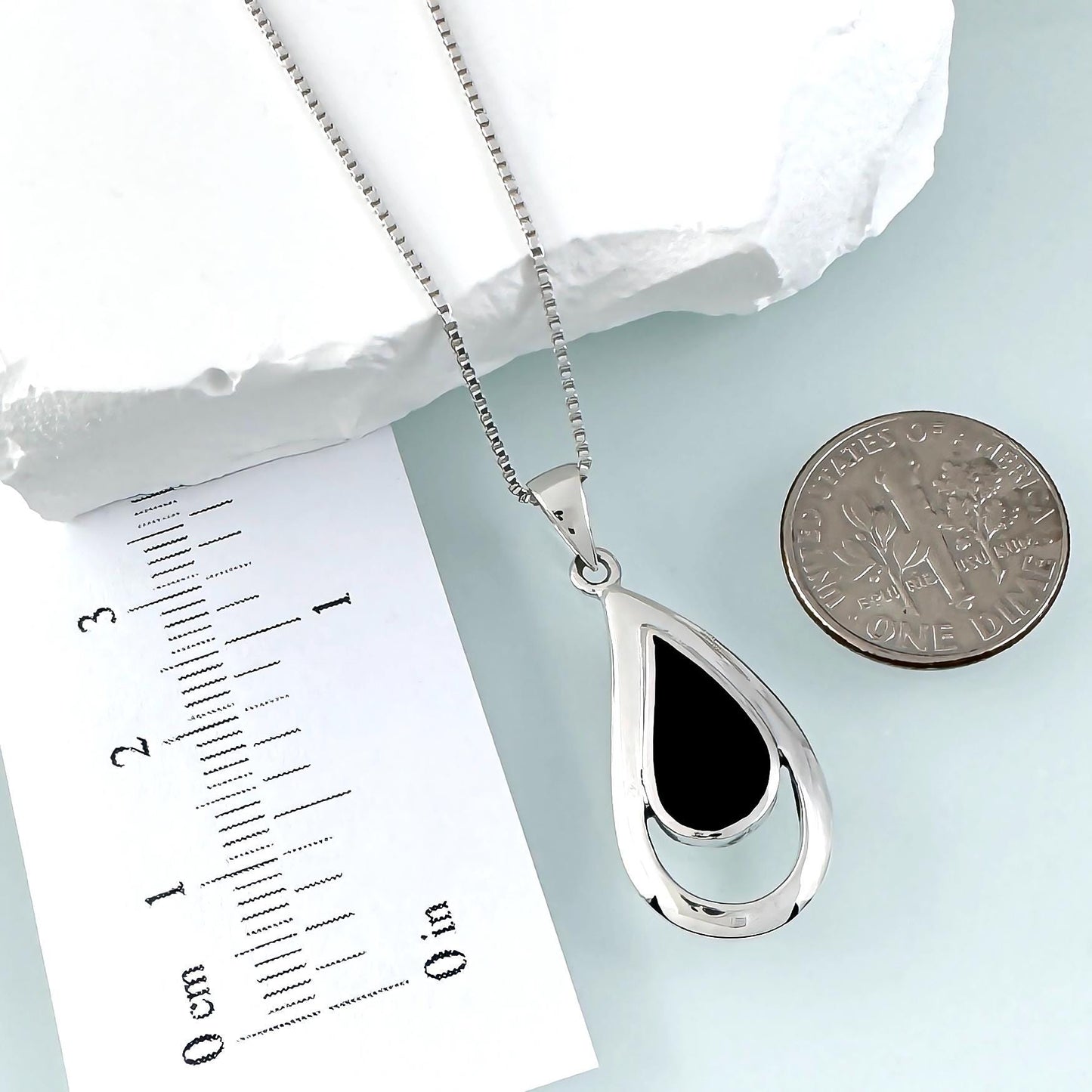 a silver necklace with a pendant, a ruler for scale, and a coin for size comparison.