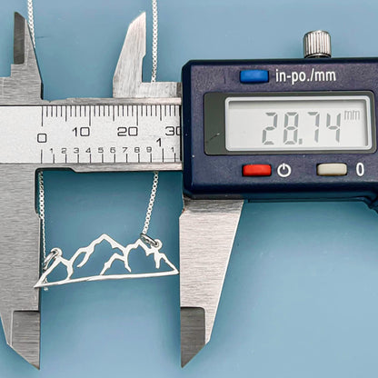A necklace with a mountain design is being measured with a ruler, and a digital caliper is also present in the image.