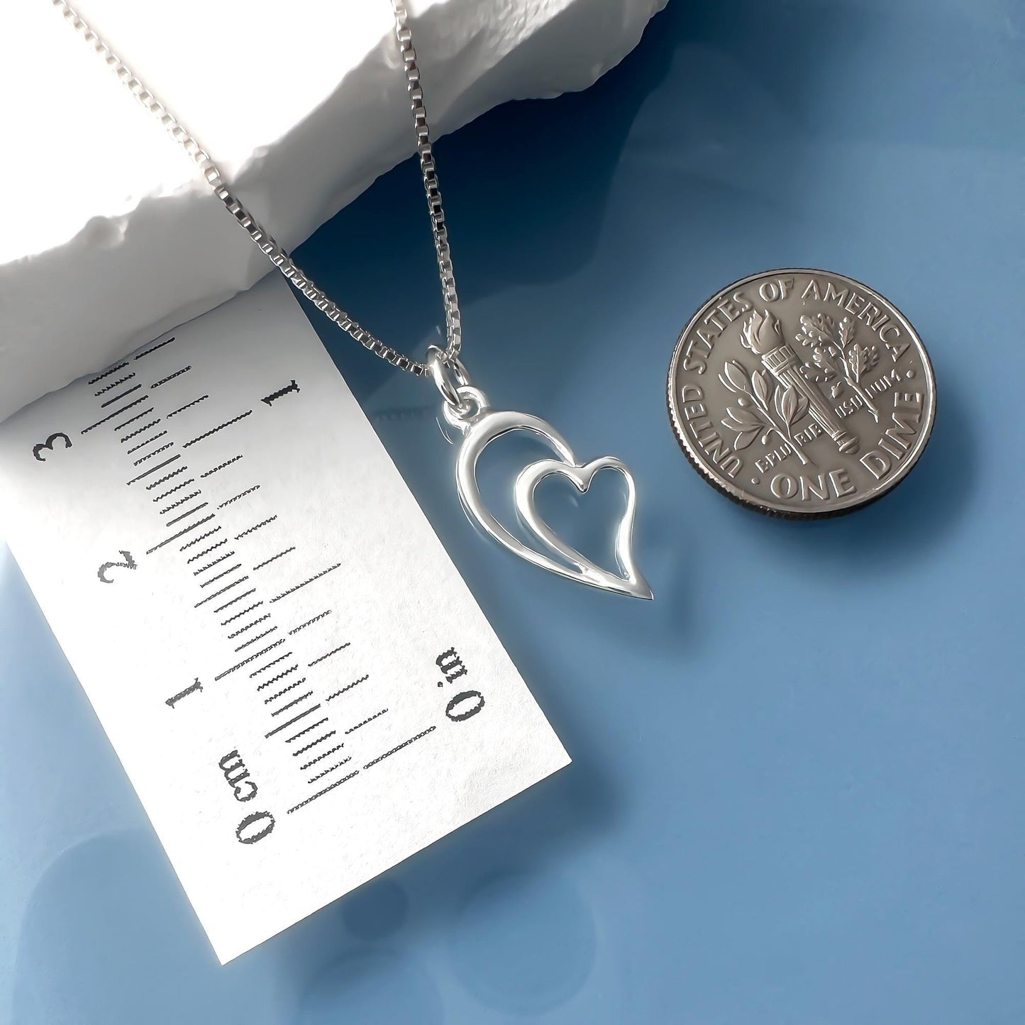 a silver heart-shaped pendant necklace and a United States one dollar coin placed on a blue surface.