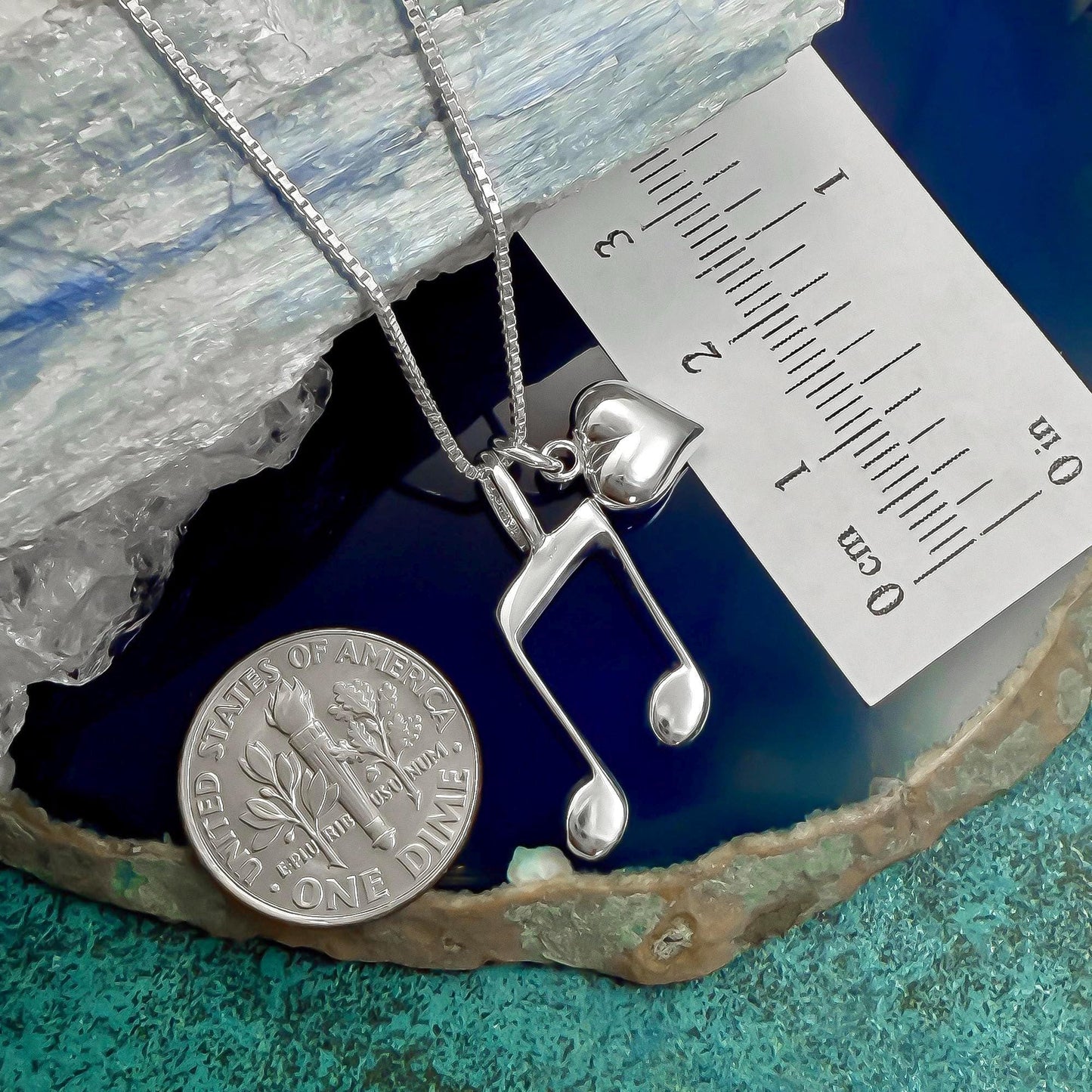 A silver necklace with a heart-shaped charm and a silver musical note pendant is displayed next to a US quarter coin, which is lying on a blue and green textured surface.