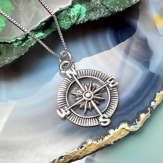 A silver compass pendant with four cardinal directions is displayed on a necklace against a colorful background.