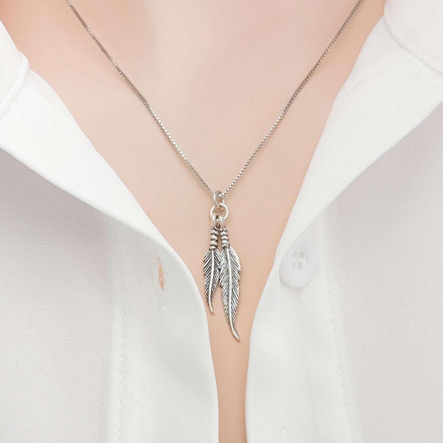 A silver necklace with a pendant featuring two feathers is shown against a white shirt.