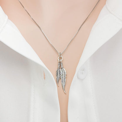 A silver necklace with a pendant featuring two feathers is shown against a white shirt.