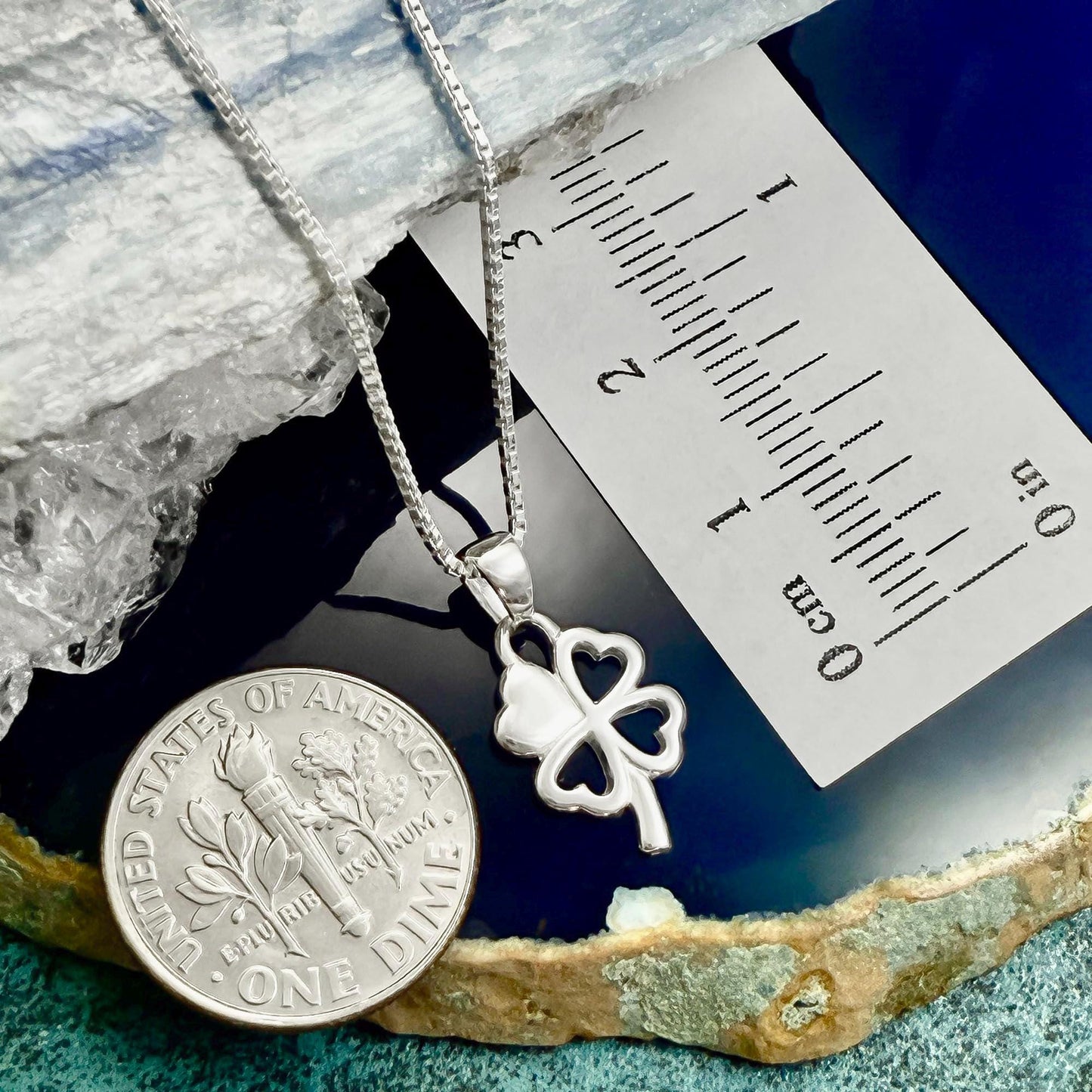 a silver-colored necklace with a four-leaf clover pendant, placed next to a United States one-dollar coin. The coin is lying on a blue and white surface, and there is a ruler placed next to it, indicating the size of the necklace.