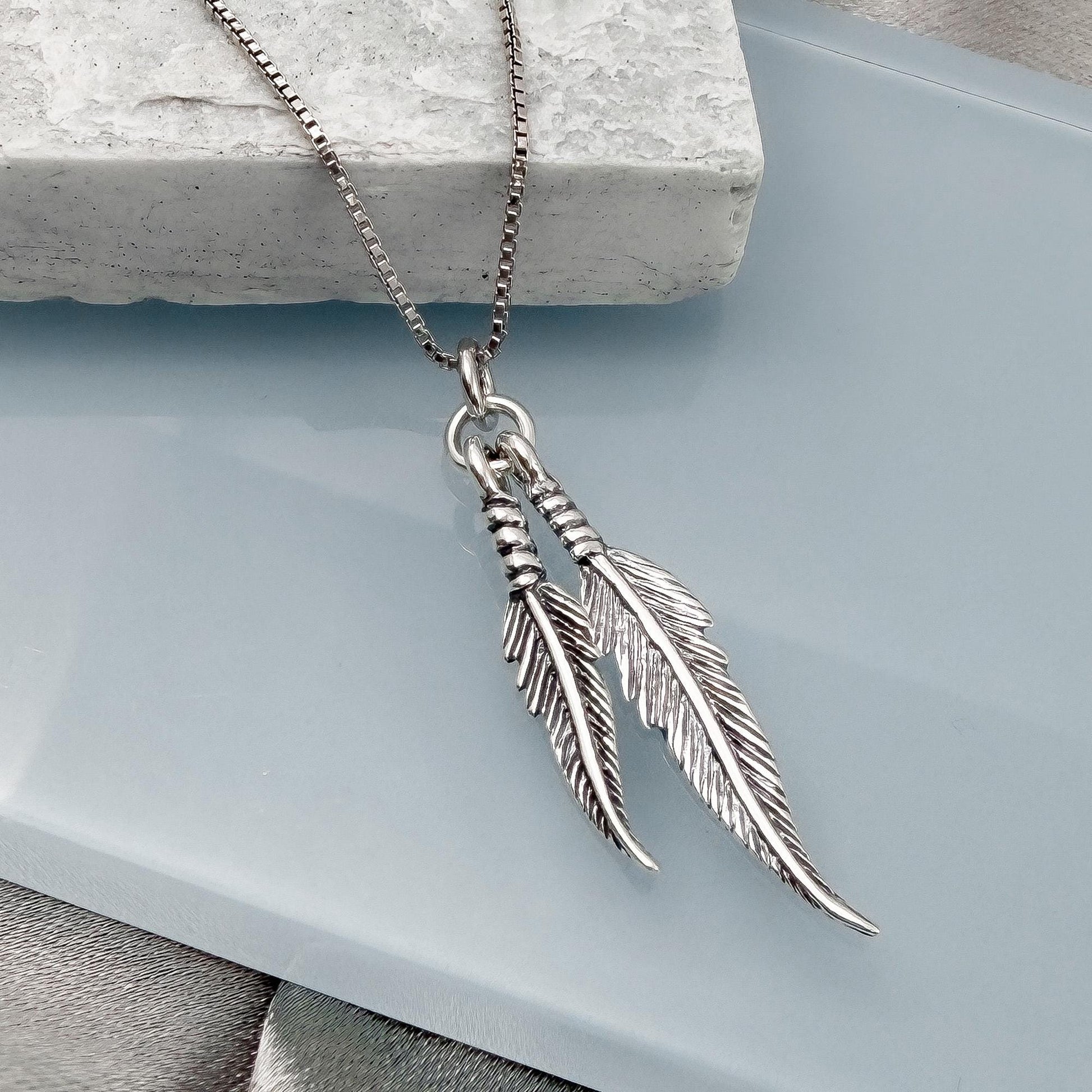 A silver-toned necklace with two feather-shaped pendants is displayed on a white surface.