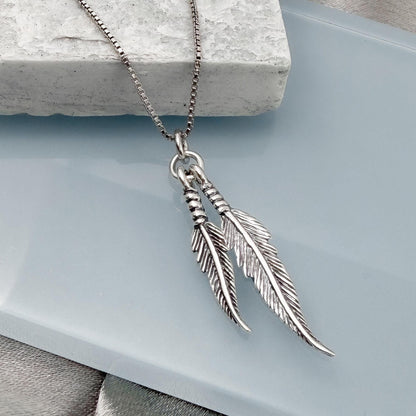 A silver-toned necklace with two feather-shaped pendants is displayed on a white surface.