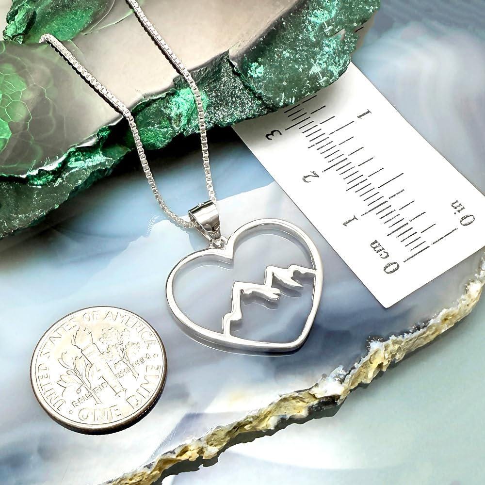 a silver heart-shaped pendant with mountain silhouettes, a silver necklace, a silver coin, and a ruler on a blue and green background.