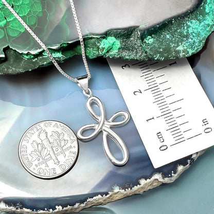 a silver cross pendant hanging from a chain, placed next to a United States one-dollar coin. The coin is lying on a blue and green surface, and there is a ruler placed next to it, indicating the size of the cross pendant.