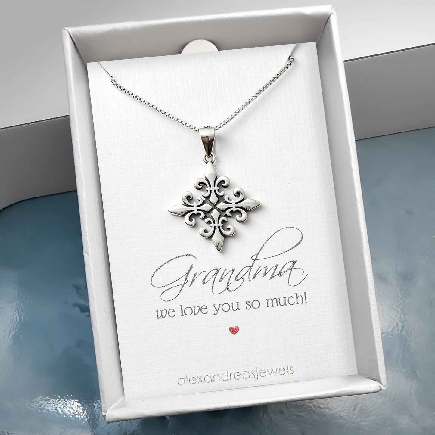 A silver necklace with a pendant is displayed in a white box, accompanied by a card that reads "Grandma we love you so much!"
