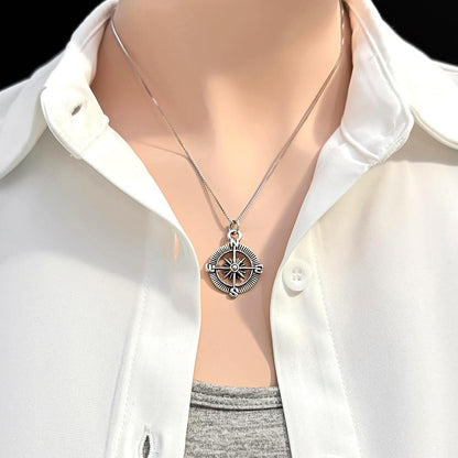 A silver compass pendant is worn on a white shirt, with the focus on the pendant and the collar of the shirt.