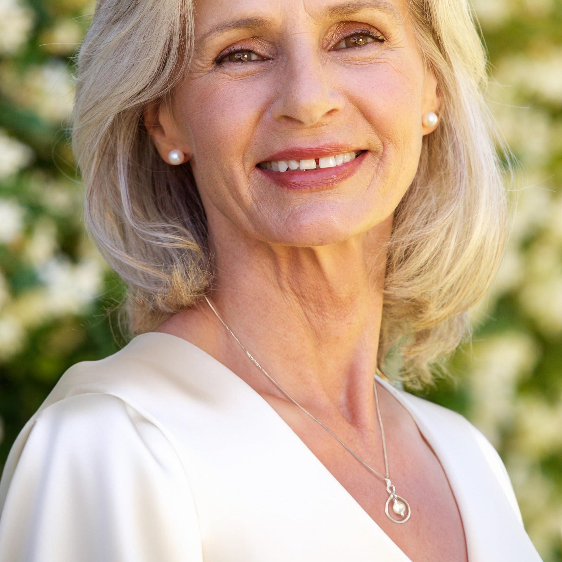 A smiling woman with blonde hair wearing a white blouse and a necklace with a pendant.