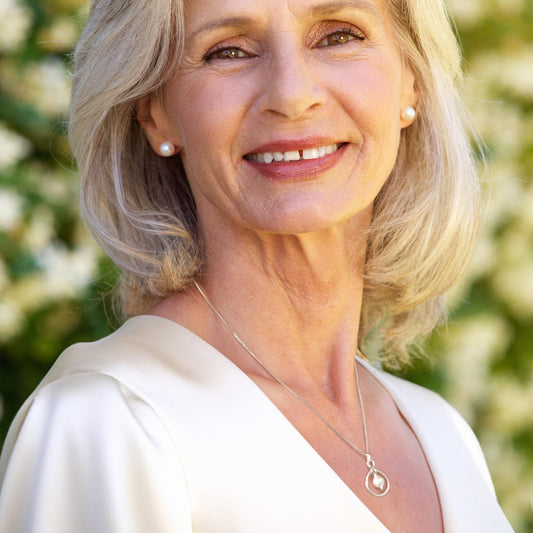 A smiling woman with blonde hair wearing a white blouse and a necklace with a pendant.