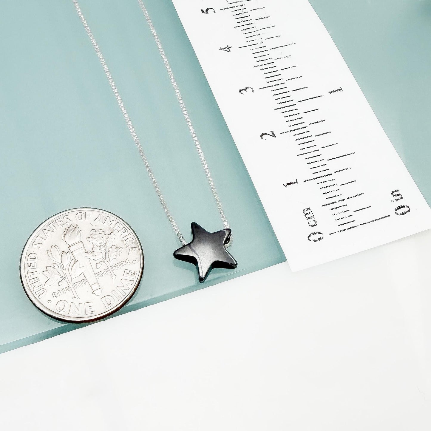 a silver necklace with a black star pendant, a silver coin, and a ruler with measurements on a light blue background.