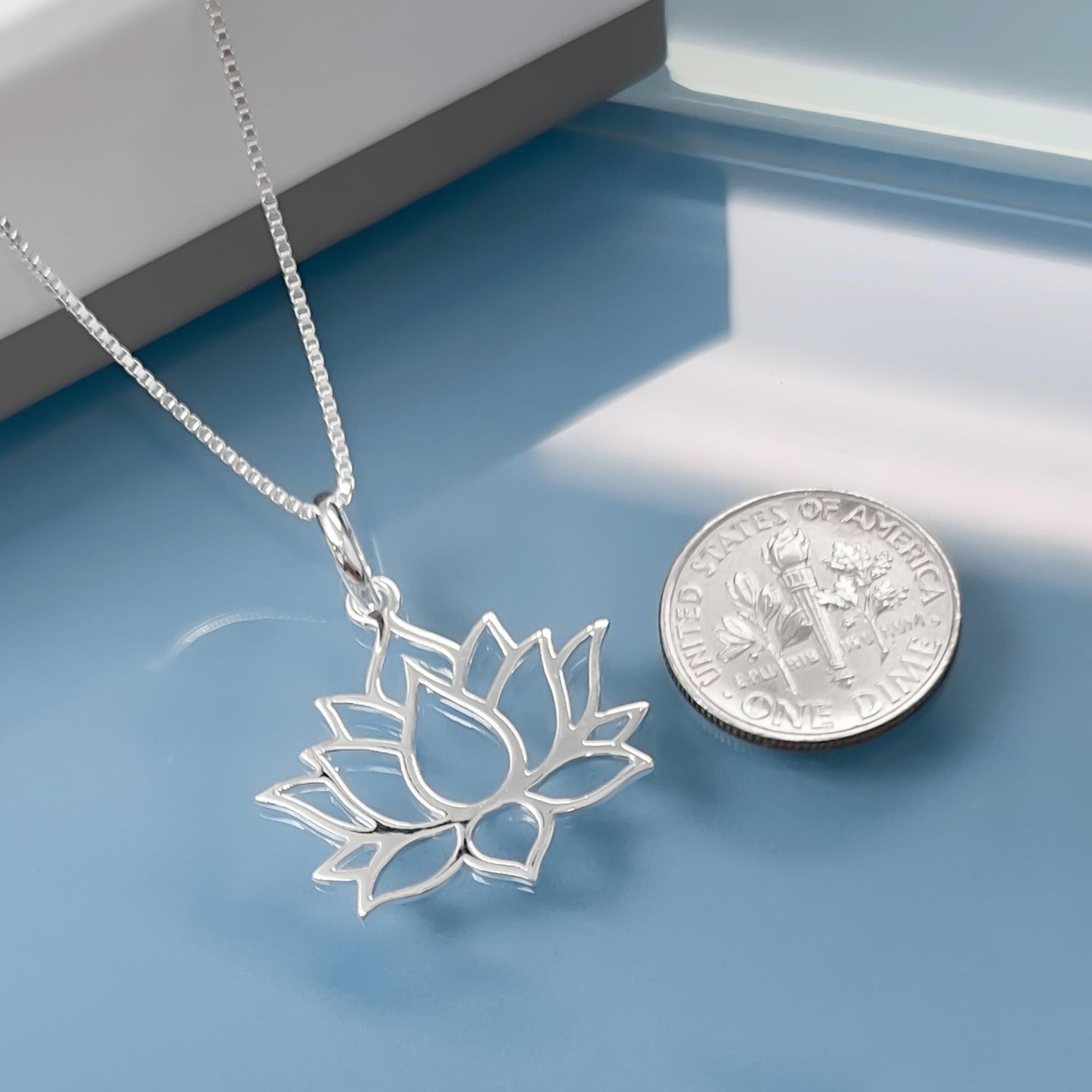 A silver necklace with a pendant in the shape of a lotus flower is displayed next to a United States one dollar coin.