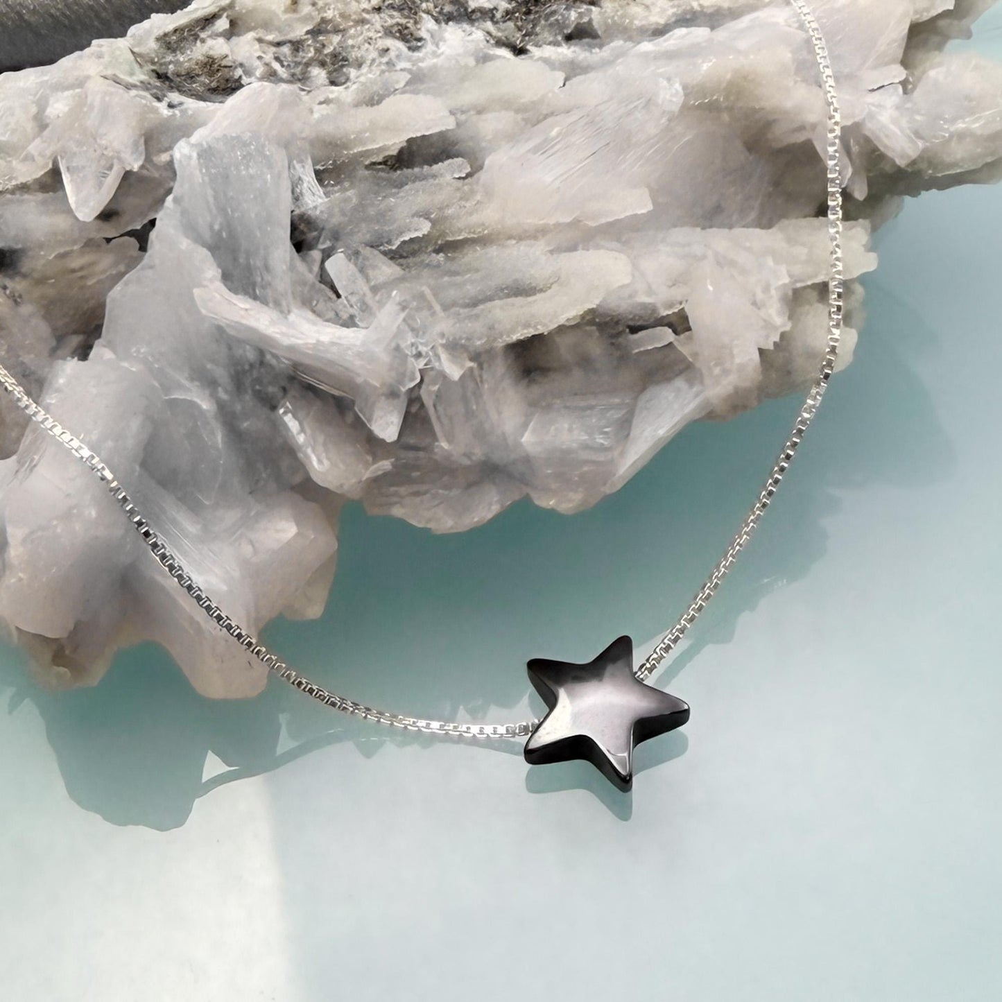 A silver necklace with a black star pendant is displayed against a backdrop of white crystals.