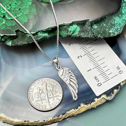 a silver necklace with a wing pendant, a United States quarter, and a ruler for size comparison.