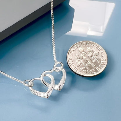 A silver necklace with two heart-shaped pendants is placed next to a United States one dollar coin on a blue surface.