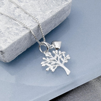 A silver tree pendant necklace with a heart charm is displayed on a gray stone surface.