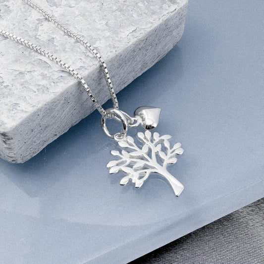 A silver tree pendant necklace with a heart charm is displayed on a gray stone surface.