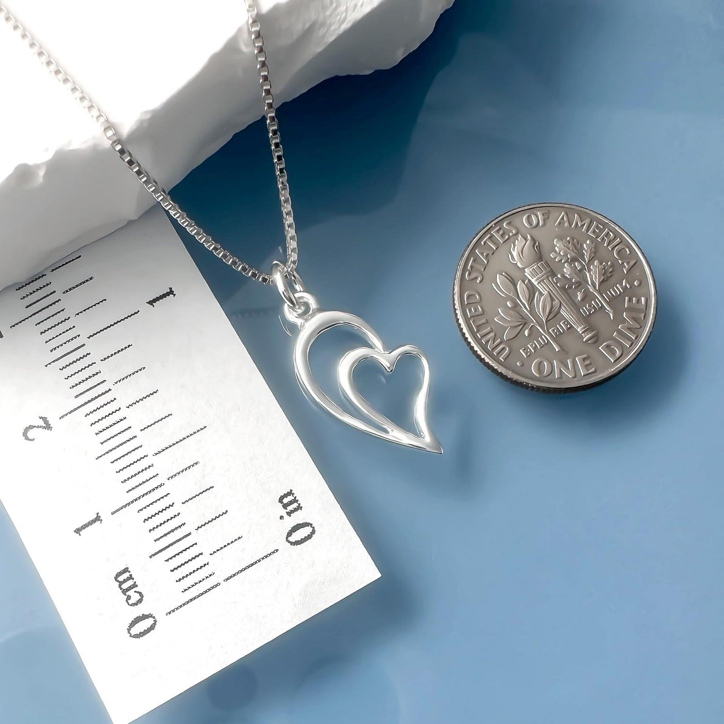 a silver heart-shaped pendant necklace and a United States one dollar coin placed on a blue surface.