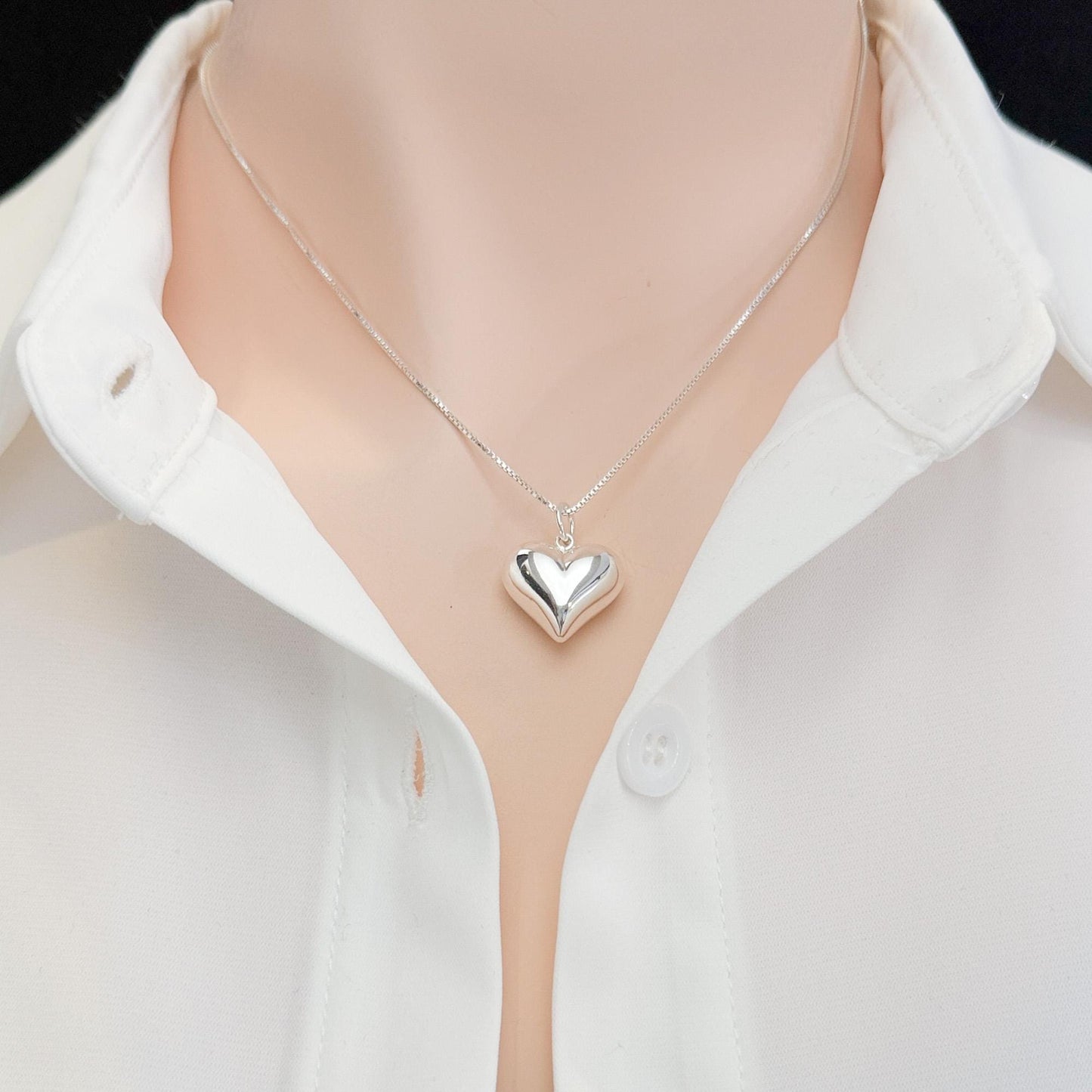 A silver heart-shaped pendant necklace is worn around a person's neck, with a white collared shirt visible underneath.