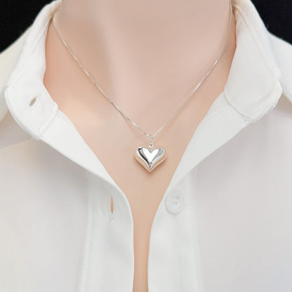 A silver heart-shaped pendant necklace is worn around a person's neck, with a white collared shirt visible underneath.