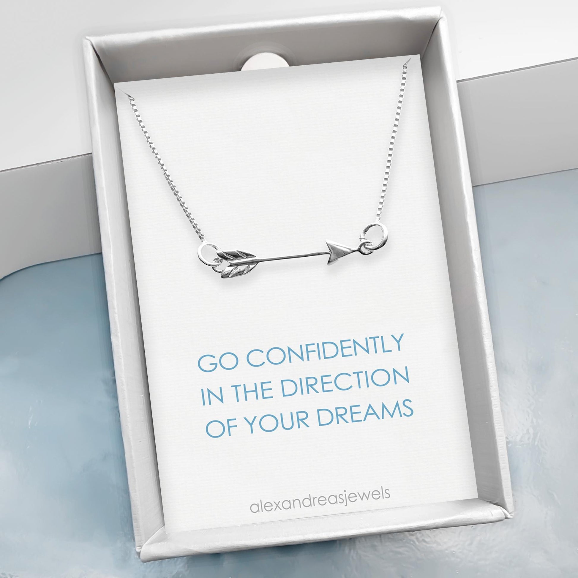 A silver necklace with an arrow pendant is displayed in a white box with a blue background.