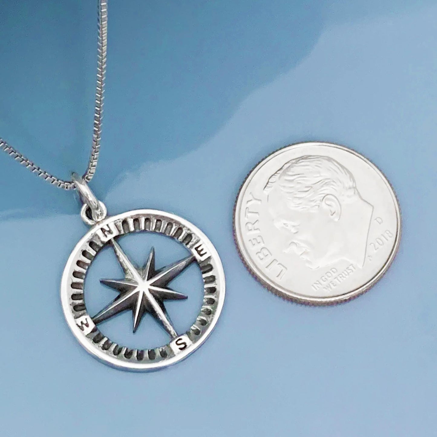 A silver-toned compass pendant necklace and a United States quarter coin are placed side by side on a blue surface.