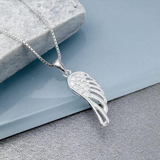 A silver pendant with a wing design is displayed on a gray surface, with a gray stone or block in the background.