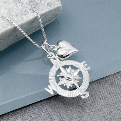 A silver compass pendant with a heart charm attached, resting on a gray surface.