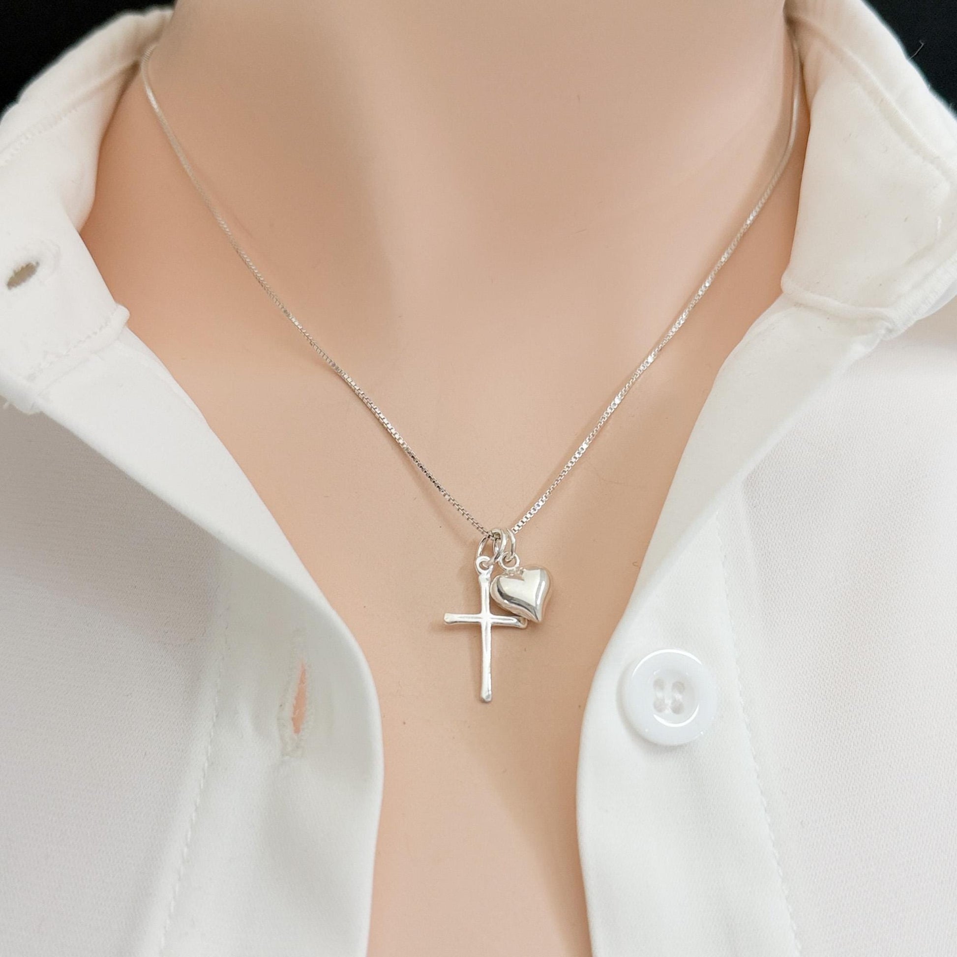 A silver cross necklace with a heart charm is worn around a person's neck.