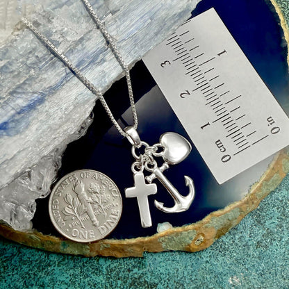 A silver necklace with a cross and an anchor charm is placed next to a United States quarter, which is approximately 1 dollar.