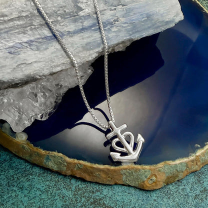 A silver necklace with a pendant featuring an anchor symbol is displayed on a blue and green textured surface.