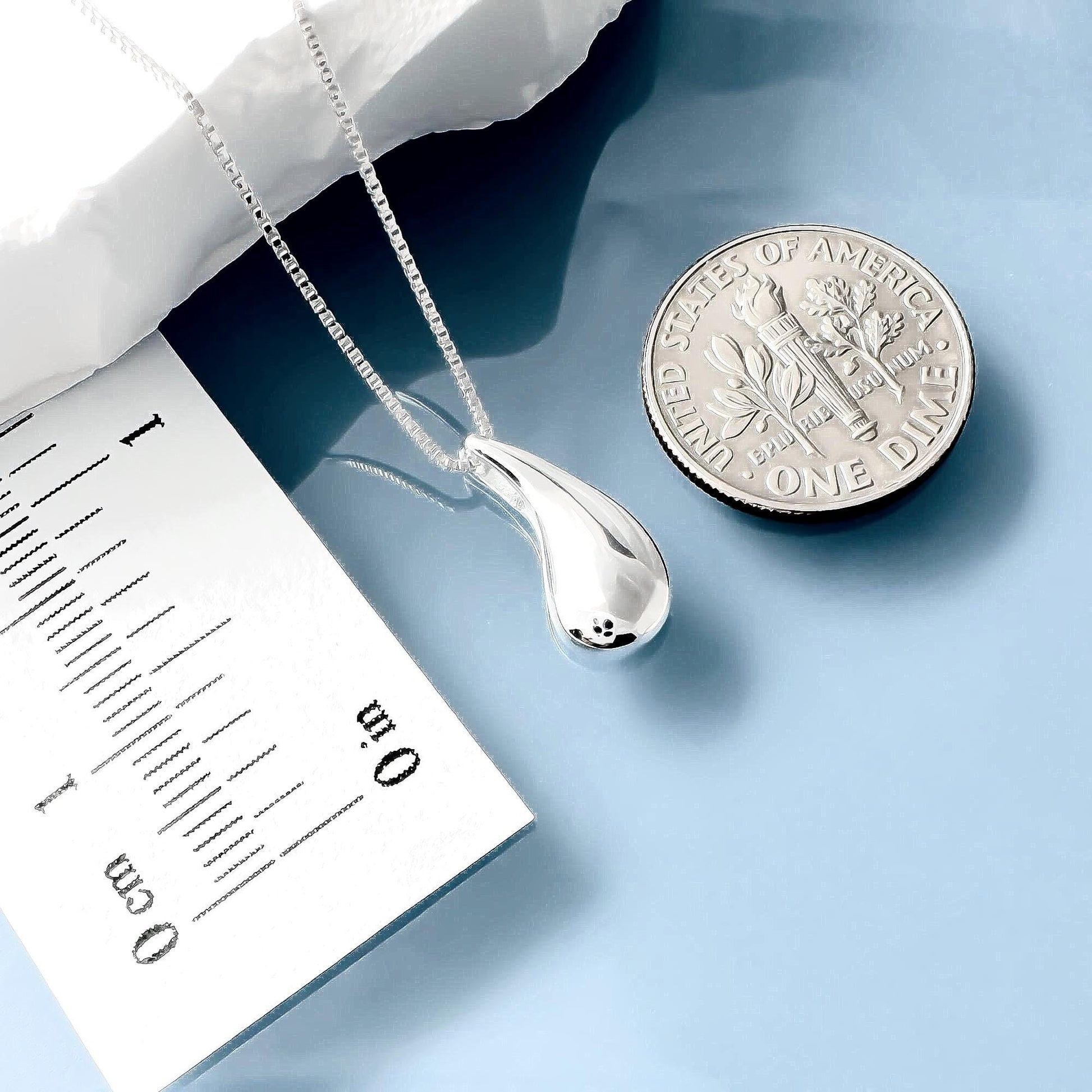 a silver necklace with a pendant, a U.S. one-dollar coin, and a paper with measurements on it, all placed on a blue surface.