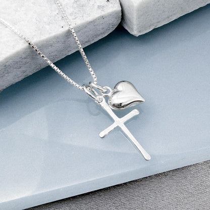 A silver cross pendant with a heart charm is displayed on a gray surface.