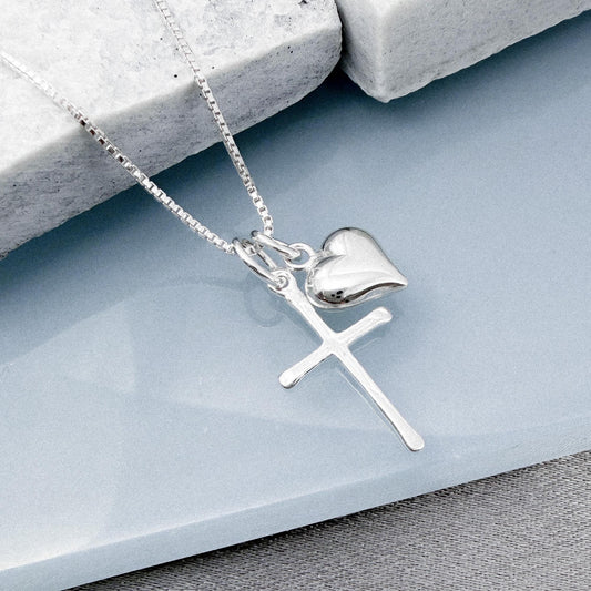A silver cross pendant with a heart charm is displayed on a gray surface.