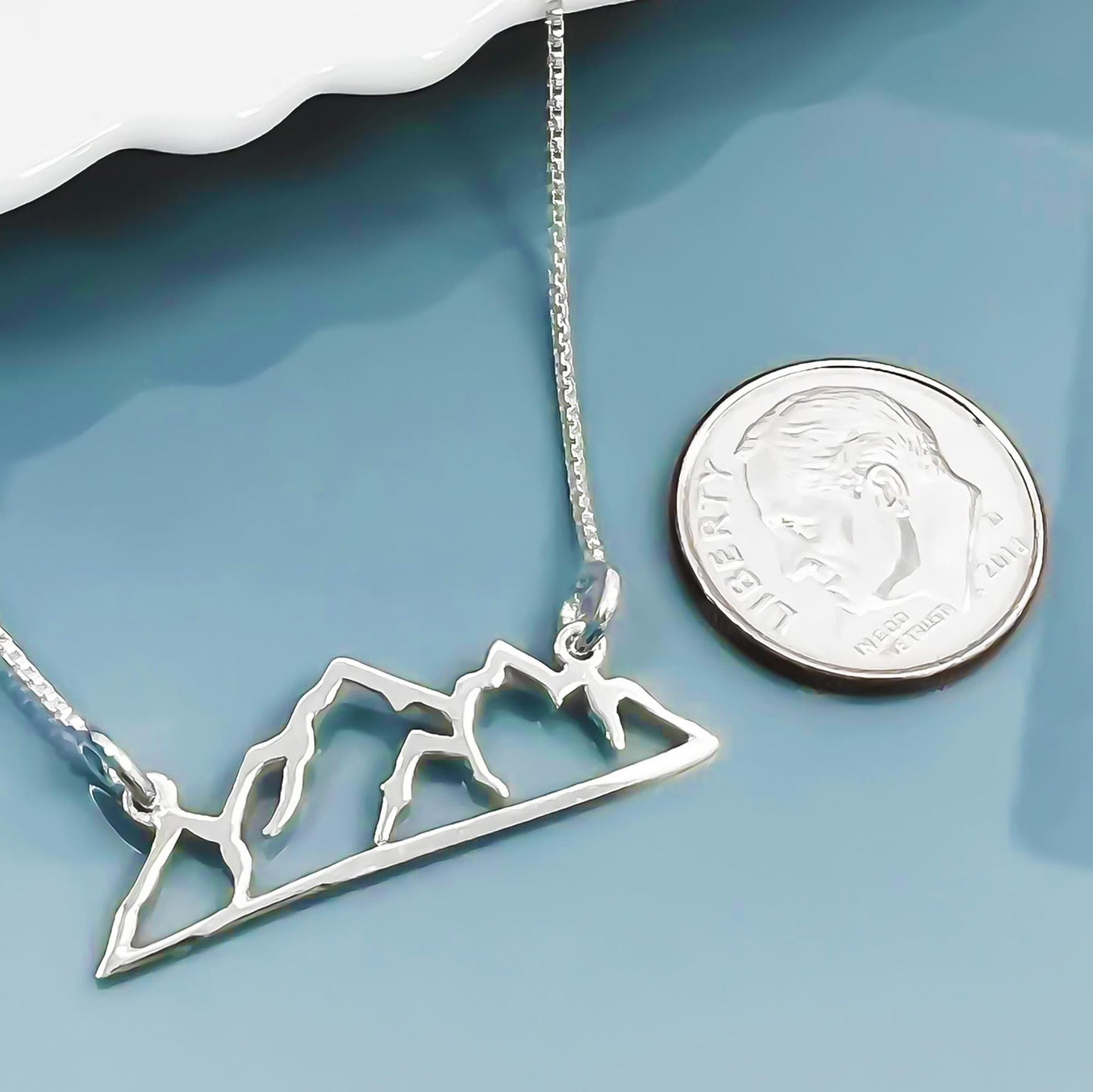 A silver necklace with a mountain silhouette pendant is placed next to a United States quarter coin on a blue surface.