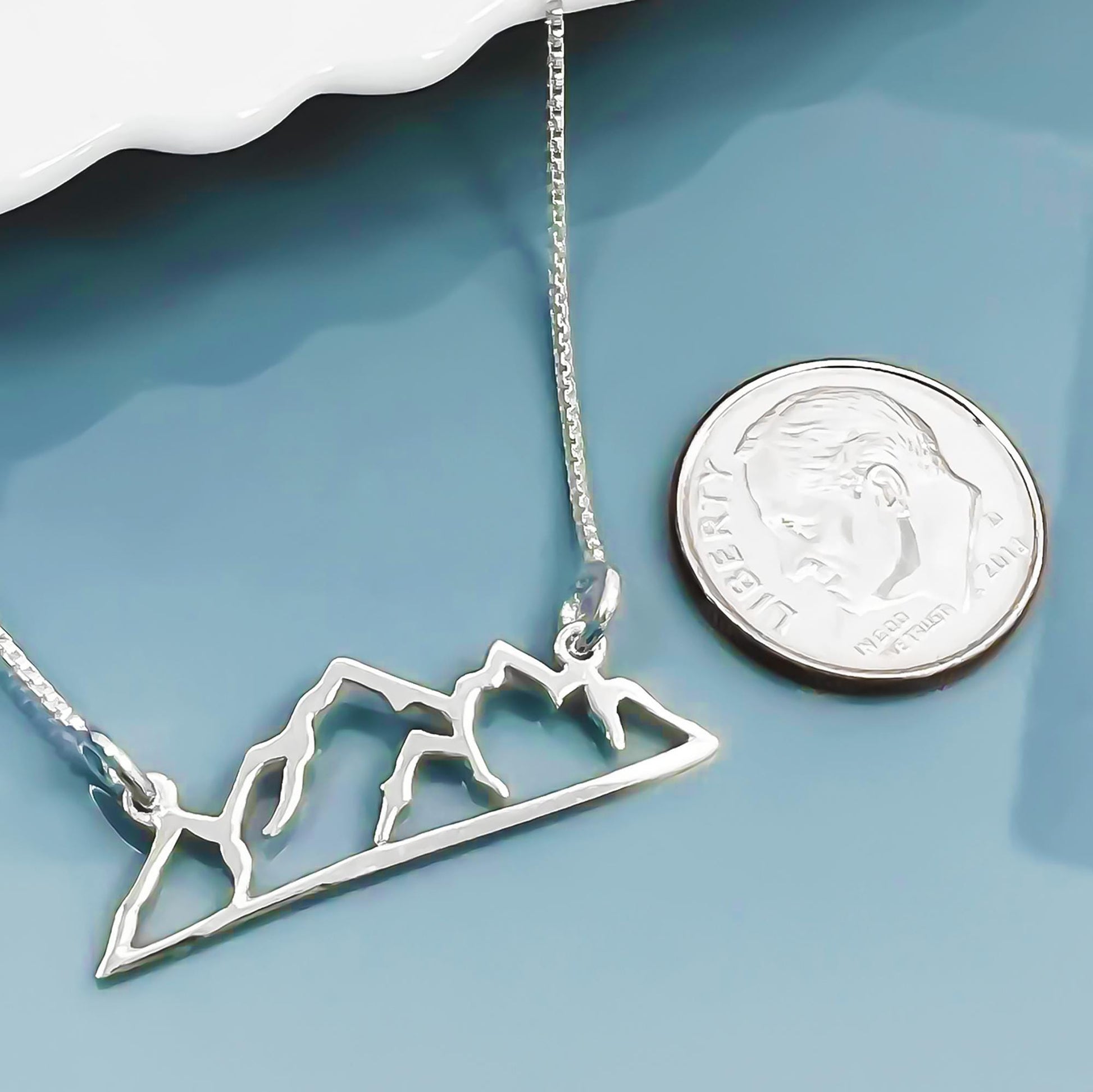 A silver necklace with a mountain silhouette pendant is placed next to a United States quarter coin on a blue surface.