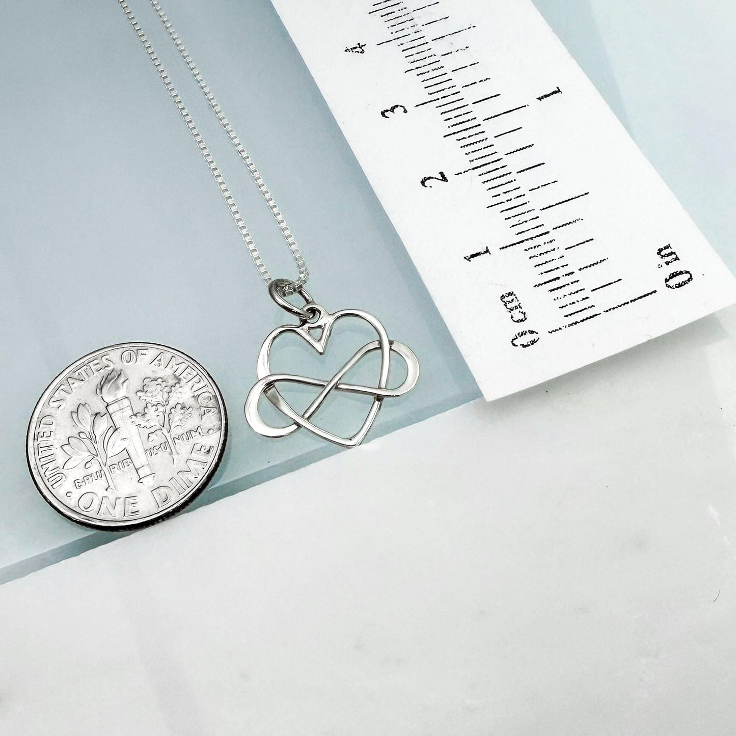 a silver necklace with a heart pendant, a silver coin, and a ruler for size comparison.