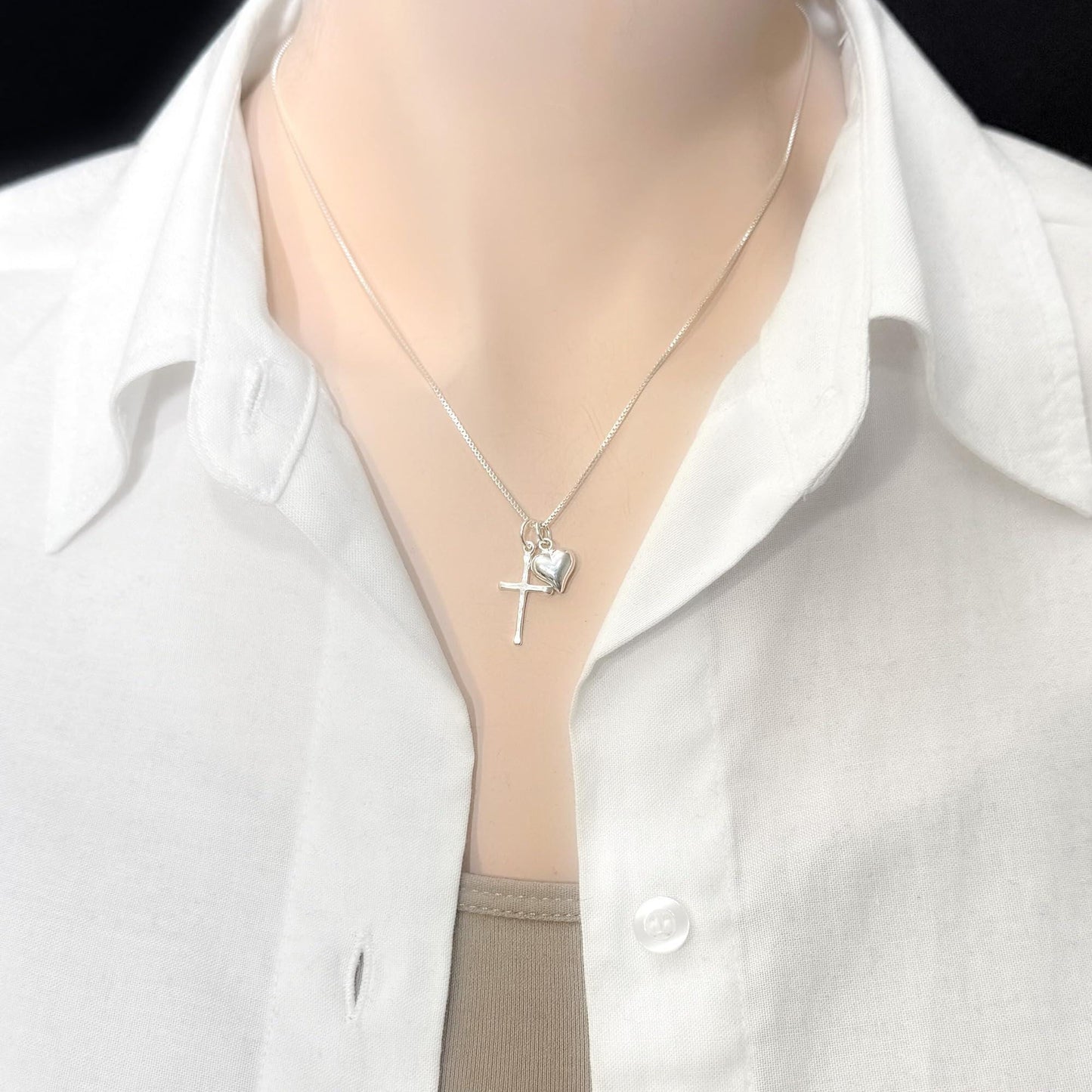 A silver necklace with a cross pendant and a small heart charm is worn around a person's neck.