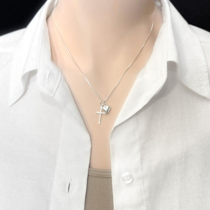 A silver necklace with a cross pendant and a small heart charm is worn around a person's neck.